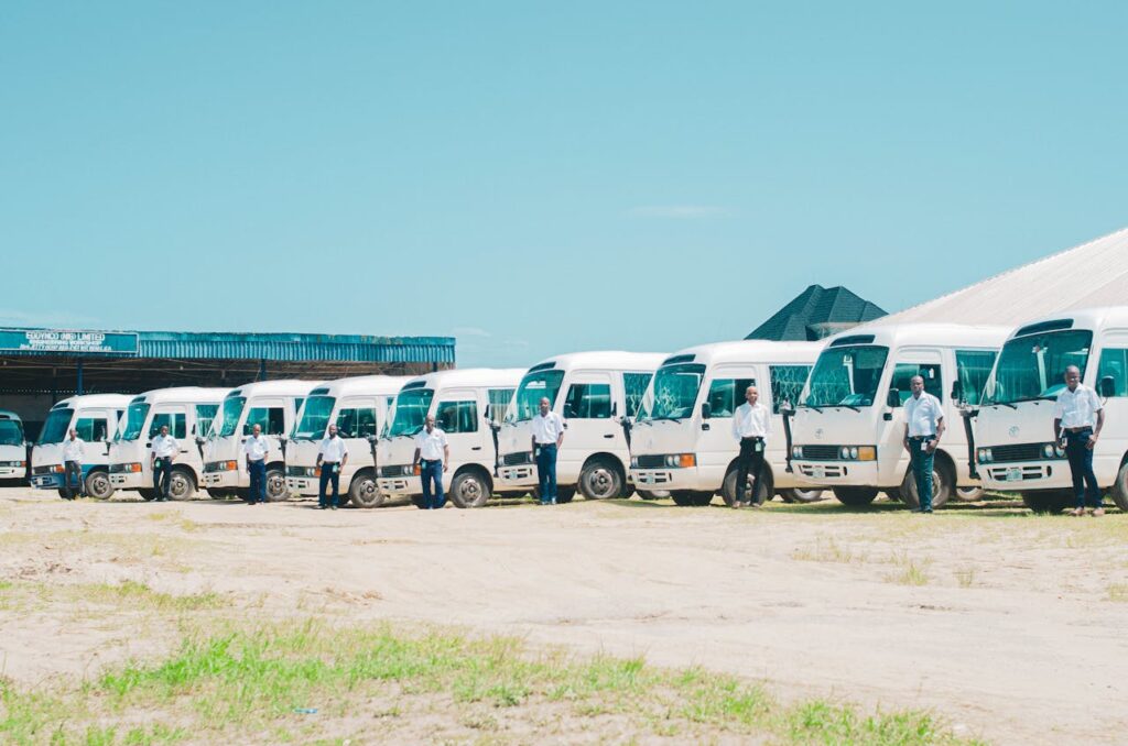 A line of minibuses with drivers standing in front, under clear skies in AK, Nigeria.