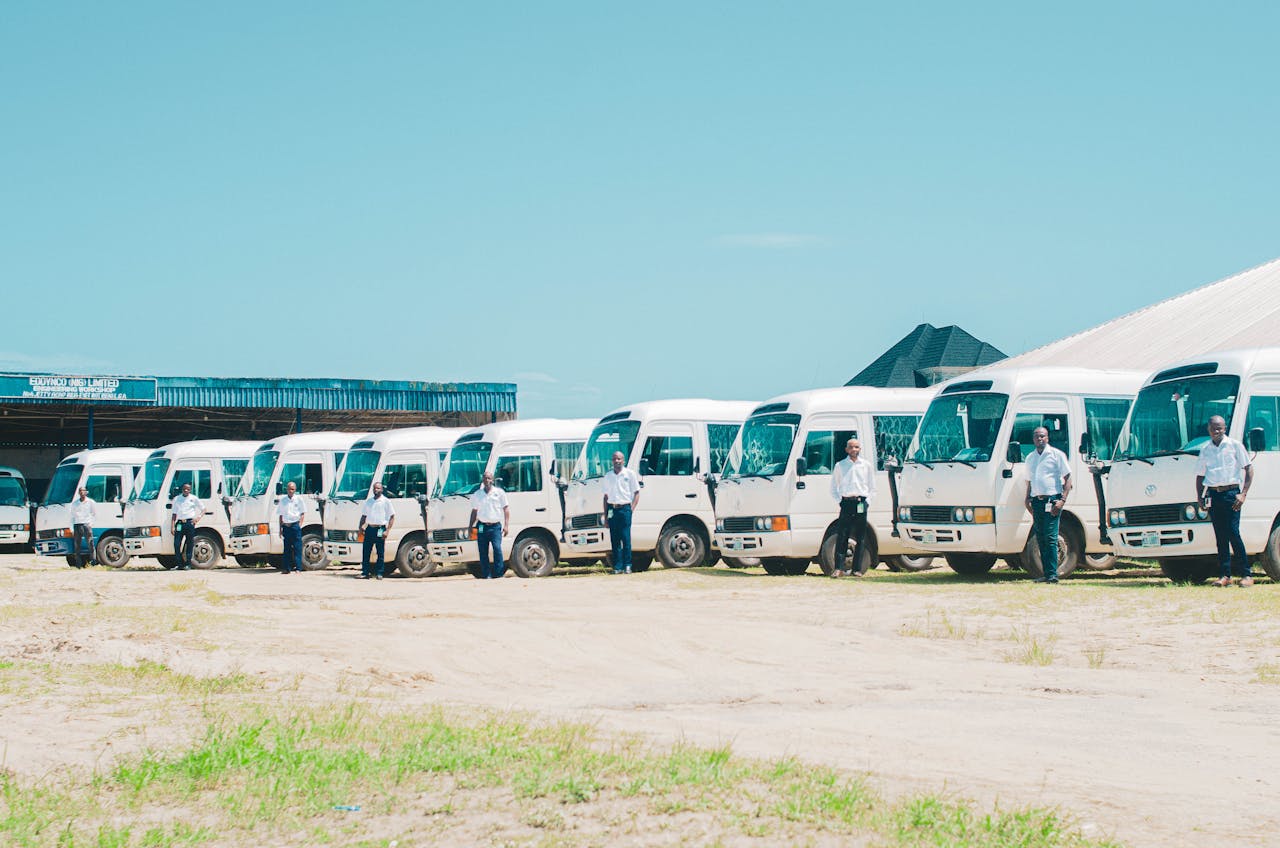 A line of minibuses with drivers standing in front, under clear skies in AK, Nigeria.