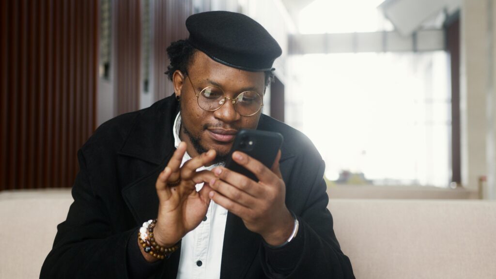 A cheerful man wearing a stylish outfit uses a smartphone while seated in a modern indoor setting.